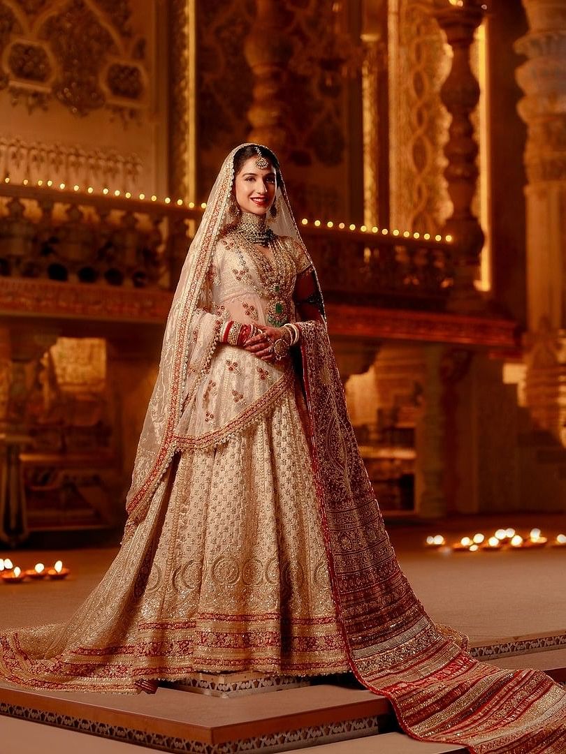 The ivory Zardozi cut-work ensemble consists of a trailing ghagra layered with a second detachable trail, a 5 meter head veil and a tissue shoulder dupatta.