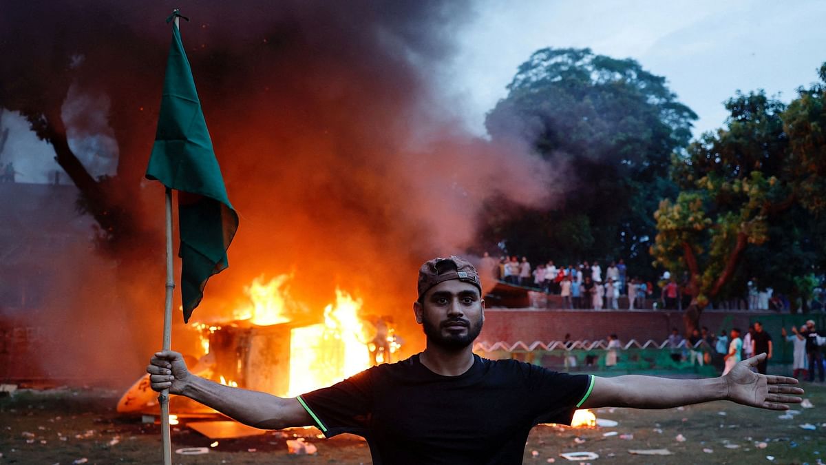 A man holding a Bangladeshi flag stands in front of a vehicle that was set on fire at the Ganabhaban, the Prime Minister's residence, after the resignation of PM Sheikh Hasina in Dhaka.