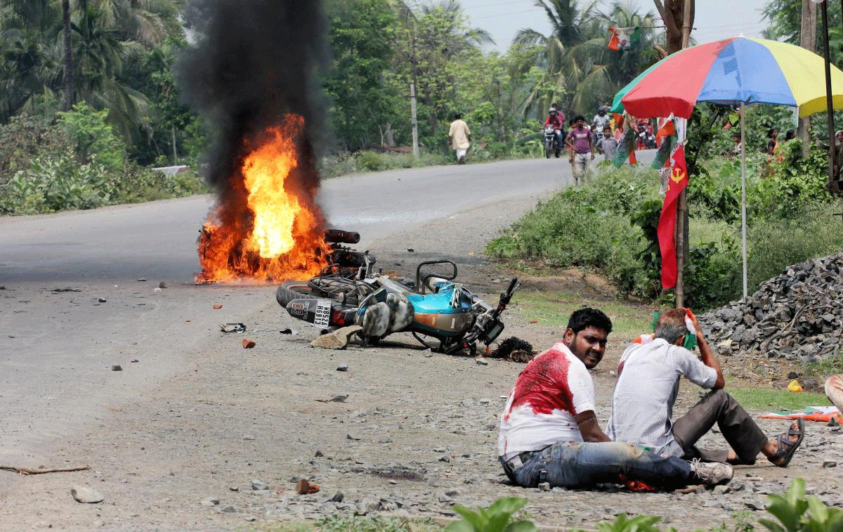 People injured in poll violence sit by the side of a road as a vehicle is set on fire by locals during Panchayat polls, in Nadia district of West Bengal on Monday. PTI Photo