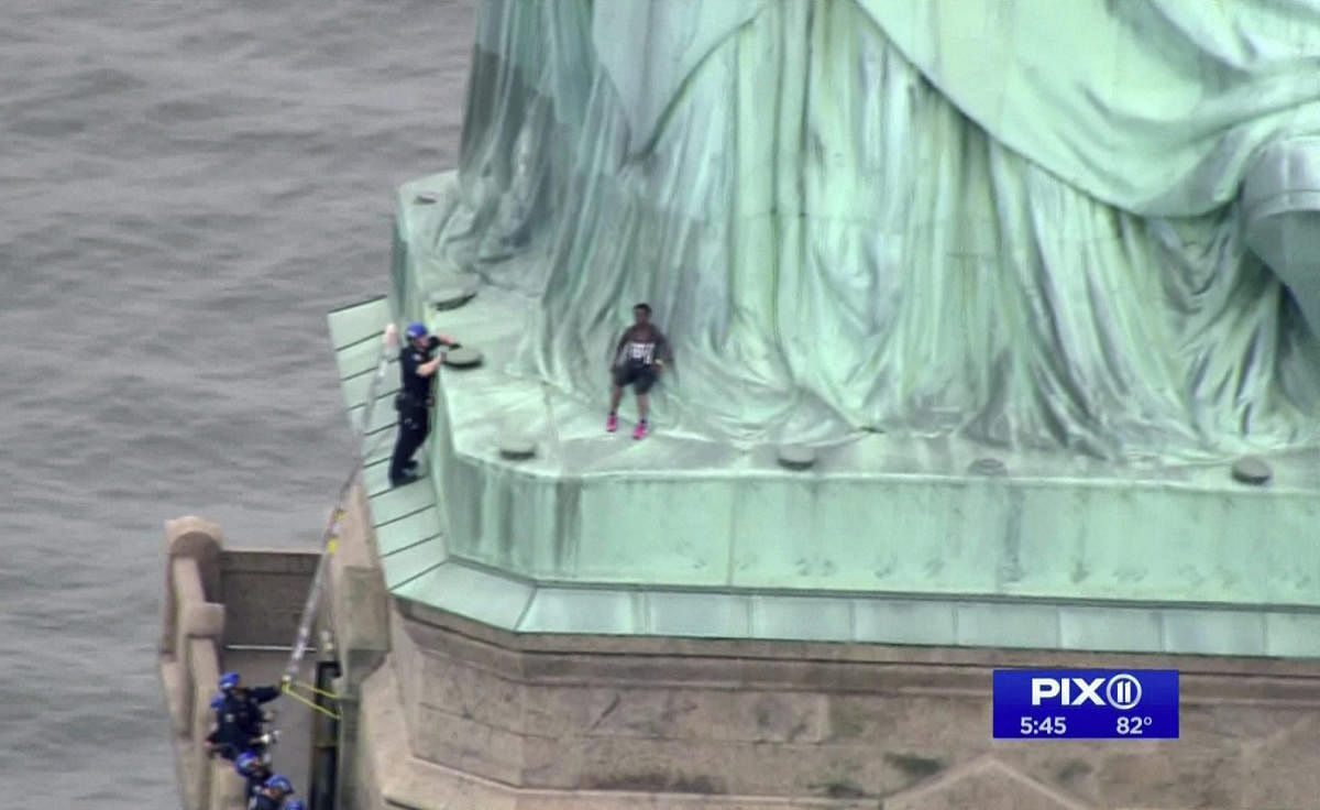 In this image made from video by PIX11, a person, center, leans against the robes of the Statue of Liberty on Liberty Island, as one of the police officers climbed up on a ladder to stand on a ledge nearby talking the climber into descending in New York, Wednesday, July 4, 2018. AP/PTI
