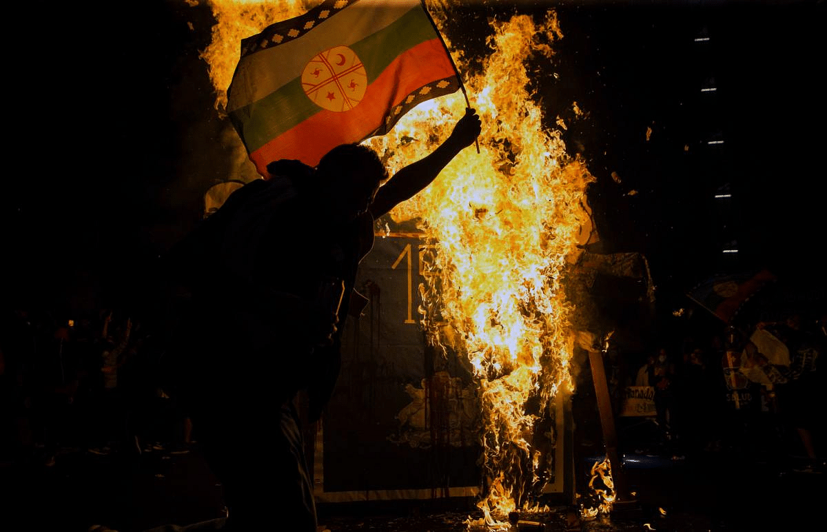 A demonstrator supporting the reform of the Chilean constitution waves a Mapuche flag in front of a sign reading Constitucion 1980 set on fire, while waiting for the referendum official results in Santiago. Credit: AFP Photo