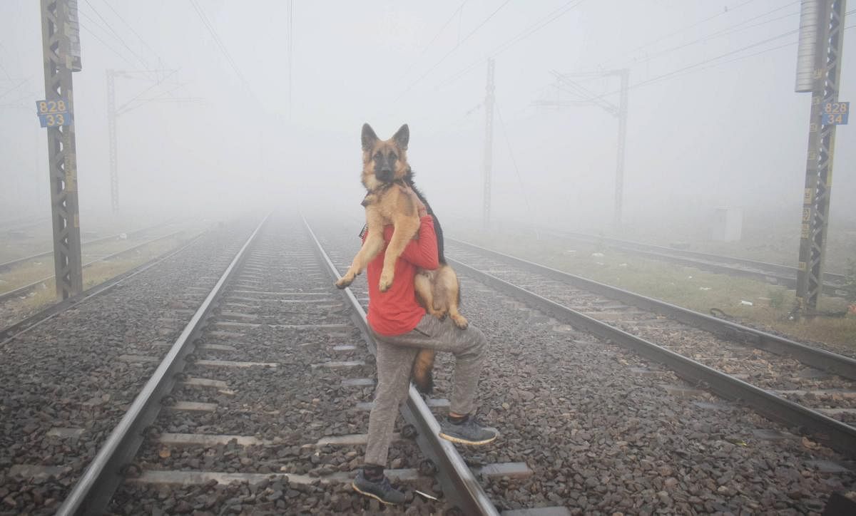 A man carrying a dog crosses a railway track, amid dense fog, during a cold morning, in Prayagraj. Credit: PTI