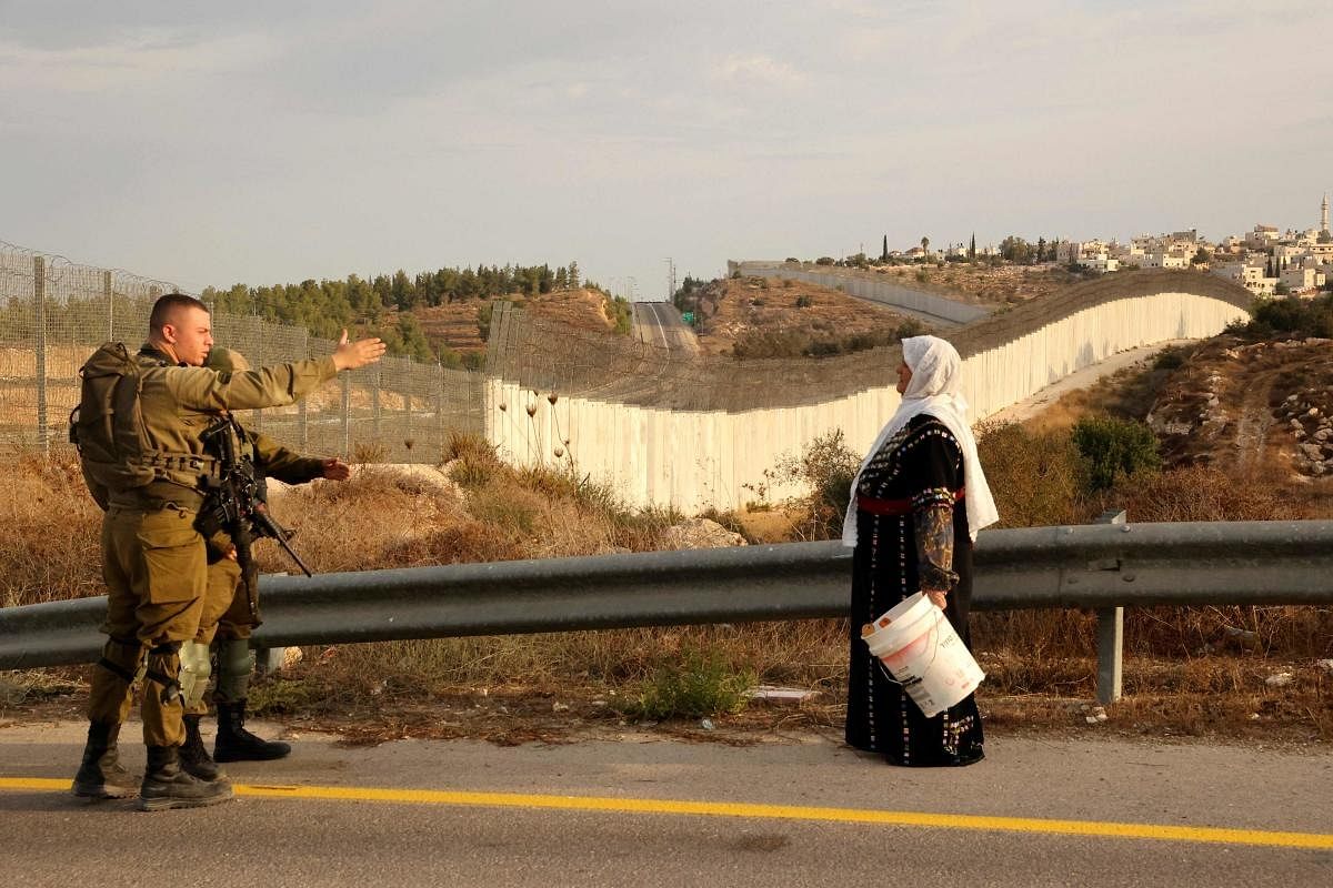 A Palestinian woman stands with others (not pictured) as they gather near an Israeli army checkpoint as they wait to reach their olives fields on the other side of Israel's separation barrier (background) after they received an special Israeli permission to harvest their olive trees. Credit: AFP Photo