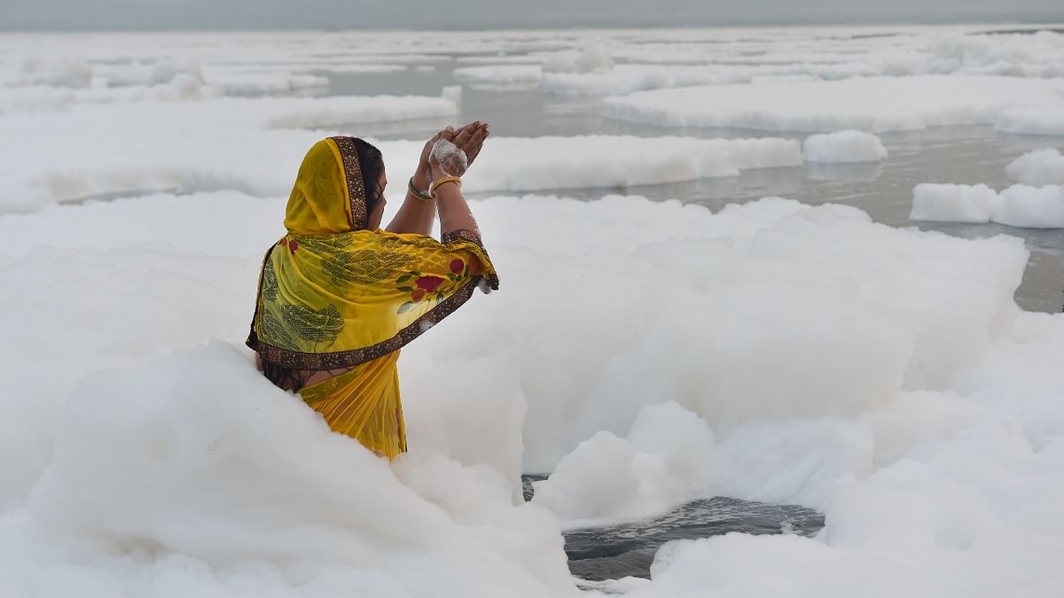 A woman offers prayers to the rising sun as toxic foam floats on the surface of polluted Yamuna river in New Delhi. Credit: PTI Photo