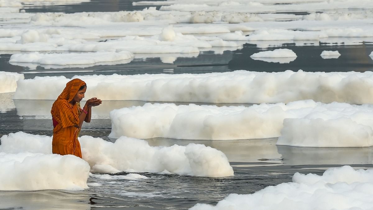 A woman devotee offers prayers to the rising sun as toxic foam floats on the surface of polluted Yamuna river during Chhath Puja celebrations, at Kalindi Kunj, in New Delhi. Credit: PTI Photo