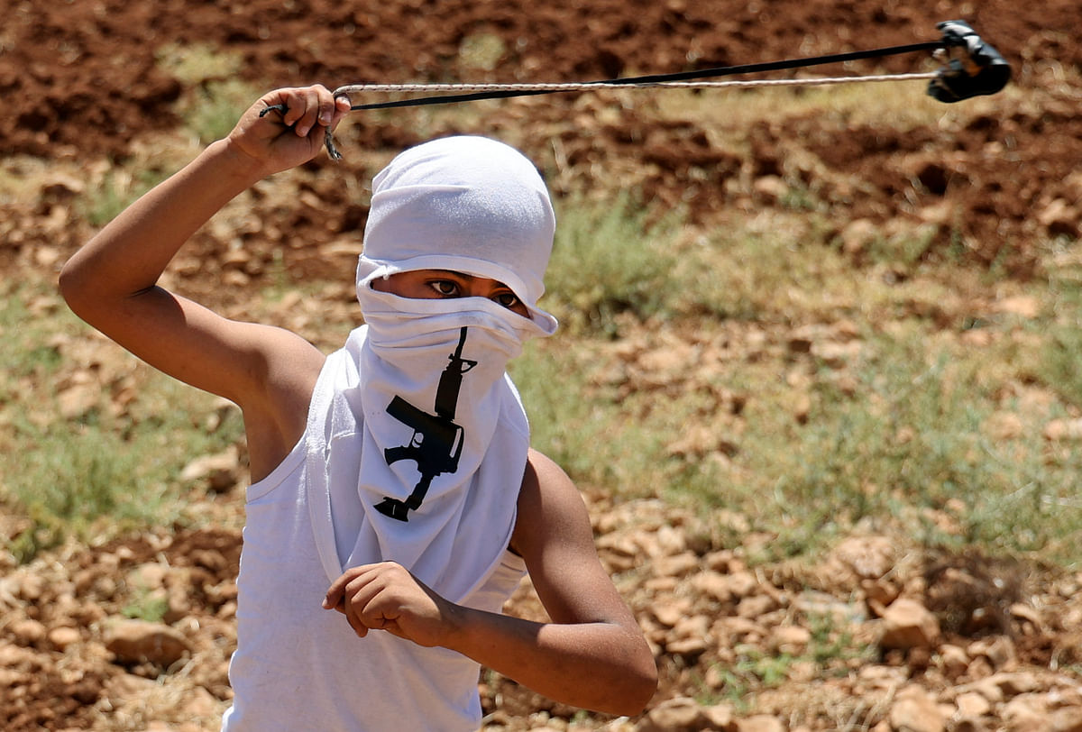 A masked Palestinian youth wearing a bandana showing an assault rifle swings a slingshot while hurling stones during clashes with Israeli forces following a demonstration against the establishment of Israeli outposts, in Beit Dajan, east of Nablus in the occupied West Bank. Credit: AFP Photo