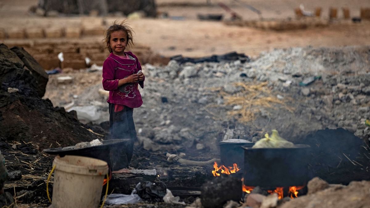 A Syrian girl stands near cooking pots placed on fire amidst the remains of asphalt at the Sahlah al-Banat camp for displaced people in the countryside of Raqa in northern Syria on November 7, 2022. Credit: AFP Photo