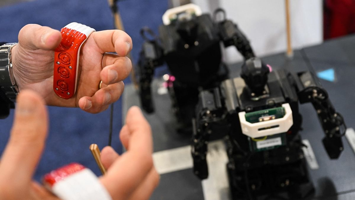 To keep up with the advent of robotics, Tactigon has developed wearables that enable gesture control for robots. The photo shows a user standing behind a small robot in the Tactigon booth at CES. The user grabs two connected joysticks and claps her hands saying