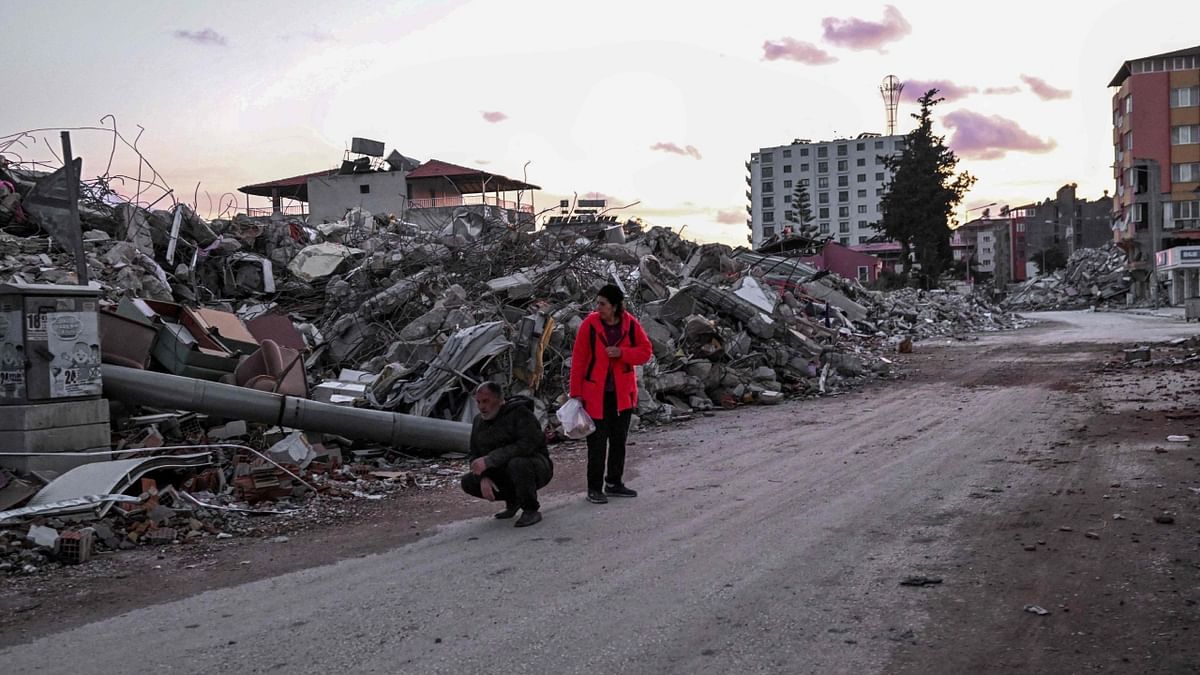 People stand on a road near the rubble of collapsed buildings, a day after a 6.4-magnitude earthquake struck the region, in the coastal city of Samandag on February 21, 2023. Credit: AFP Photo