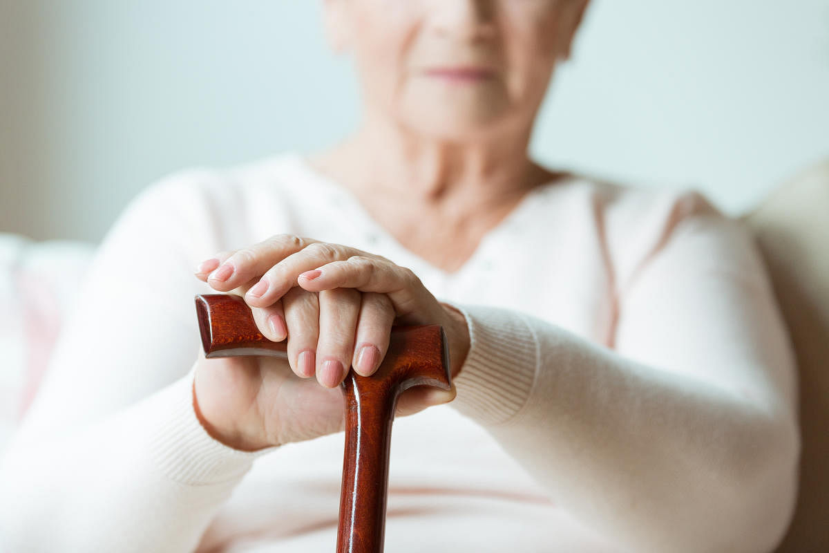 Elder holds hands on walking stick while sitting on white sofa in nursing homeParkinson's disease