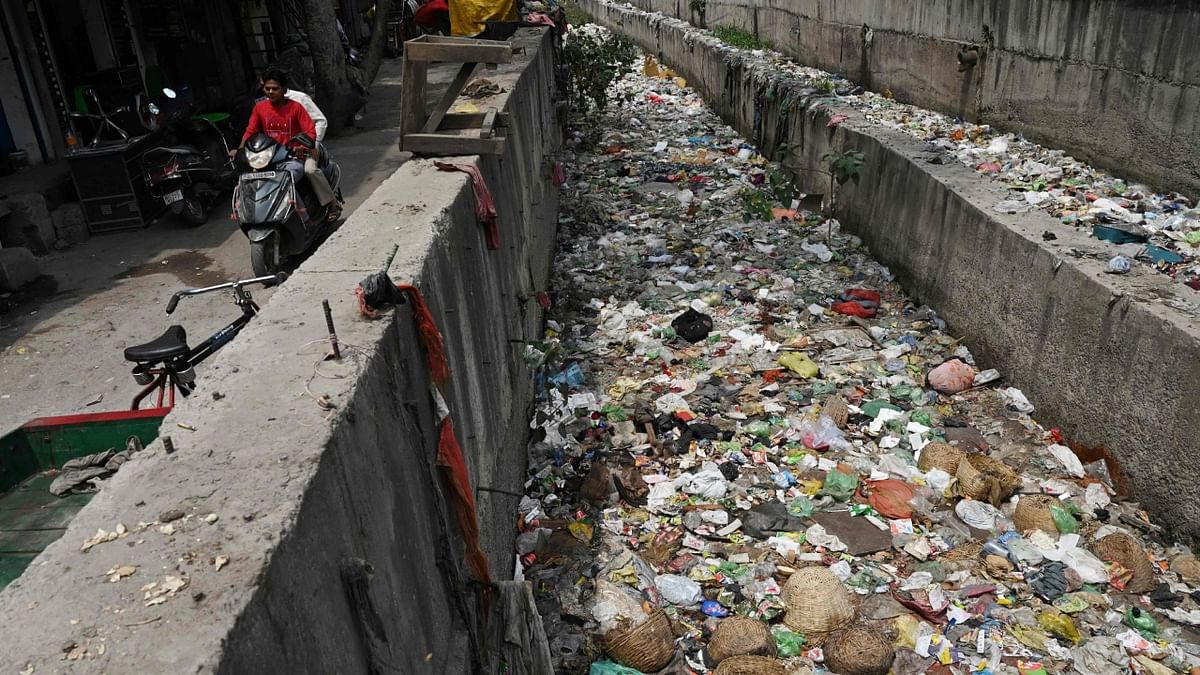 In this photograph taken on April 26, 2023, men ride a scooter along a street past a sewage canal filled with garbage at Seelampur neighbourhood in New Delhi. Credit: AFP Photo