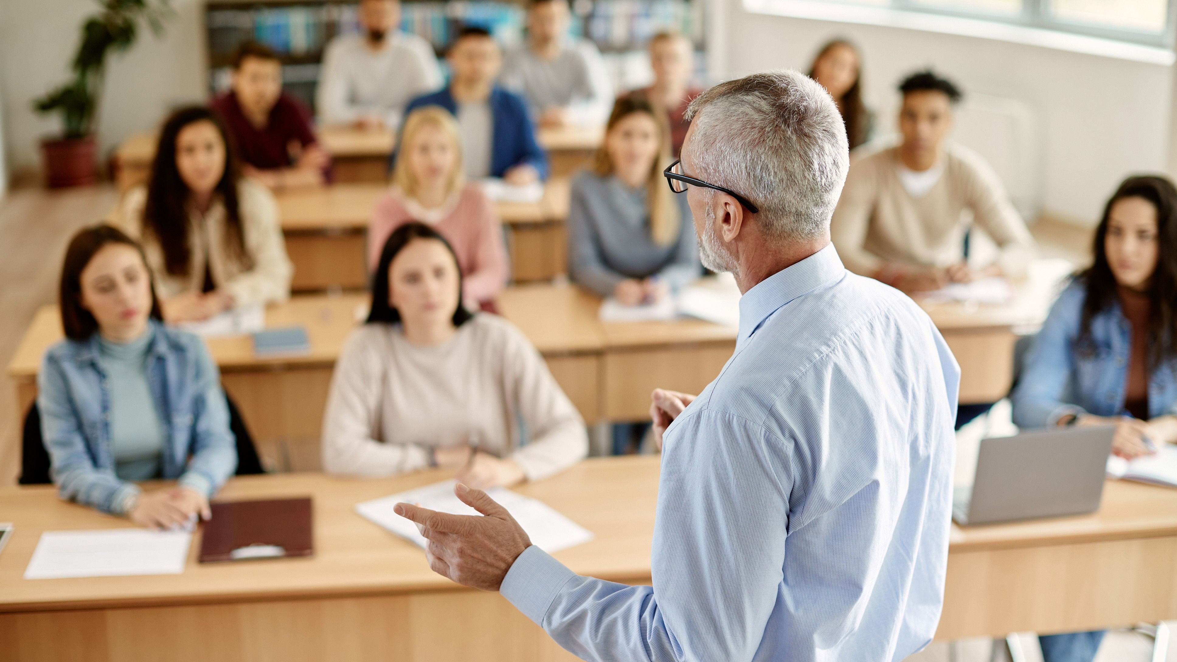 Back view of mature professor giving lecture to large group of college students in the classroom.