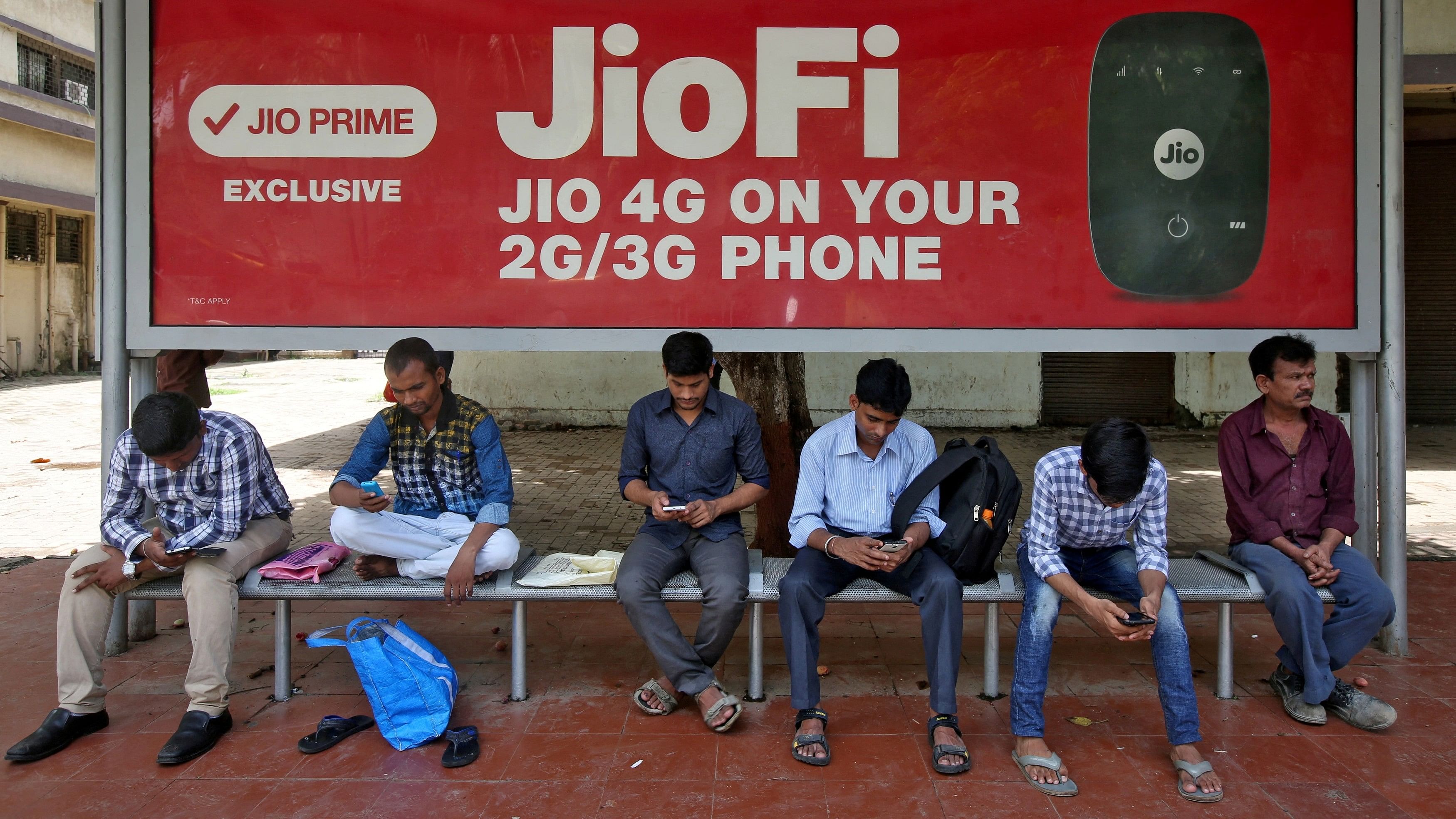 <div class="paragraphs"><p>Commuters use their mobile phones as they wait at a bus stop with an advertisement of Reliance Industries' Jio telecoms unit.</p></div>