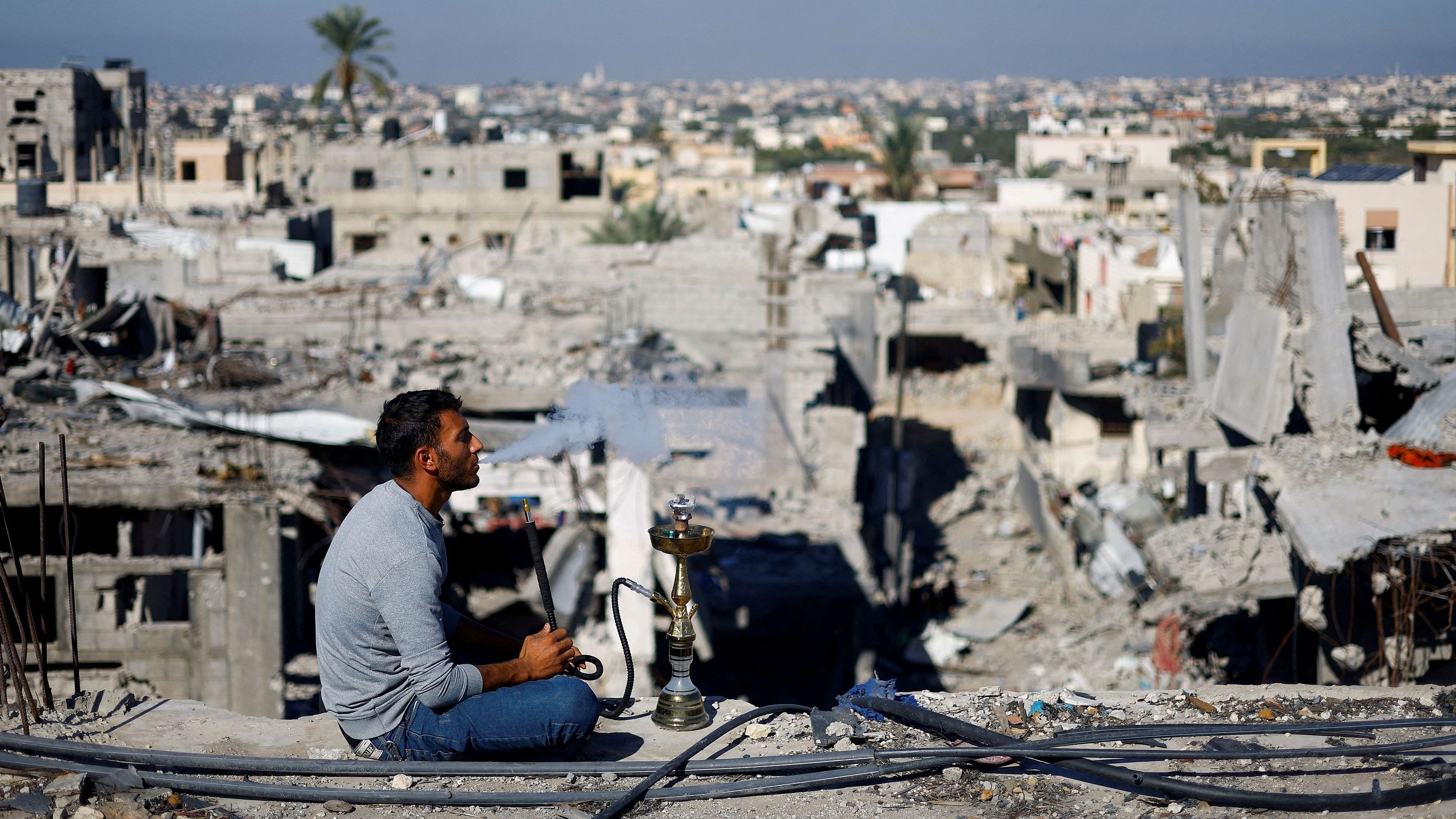 <div class="paragraphs"><p>In this file image, a Palestinian man smokes shisha as he looks at the houses destroyed in Israeli strikes.</p></div>