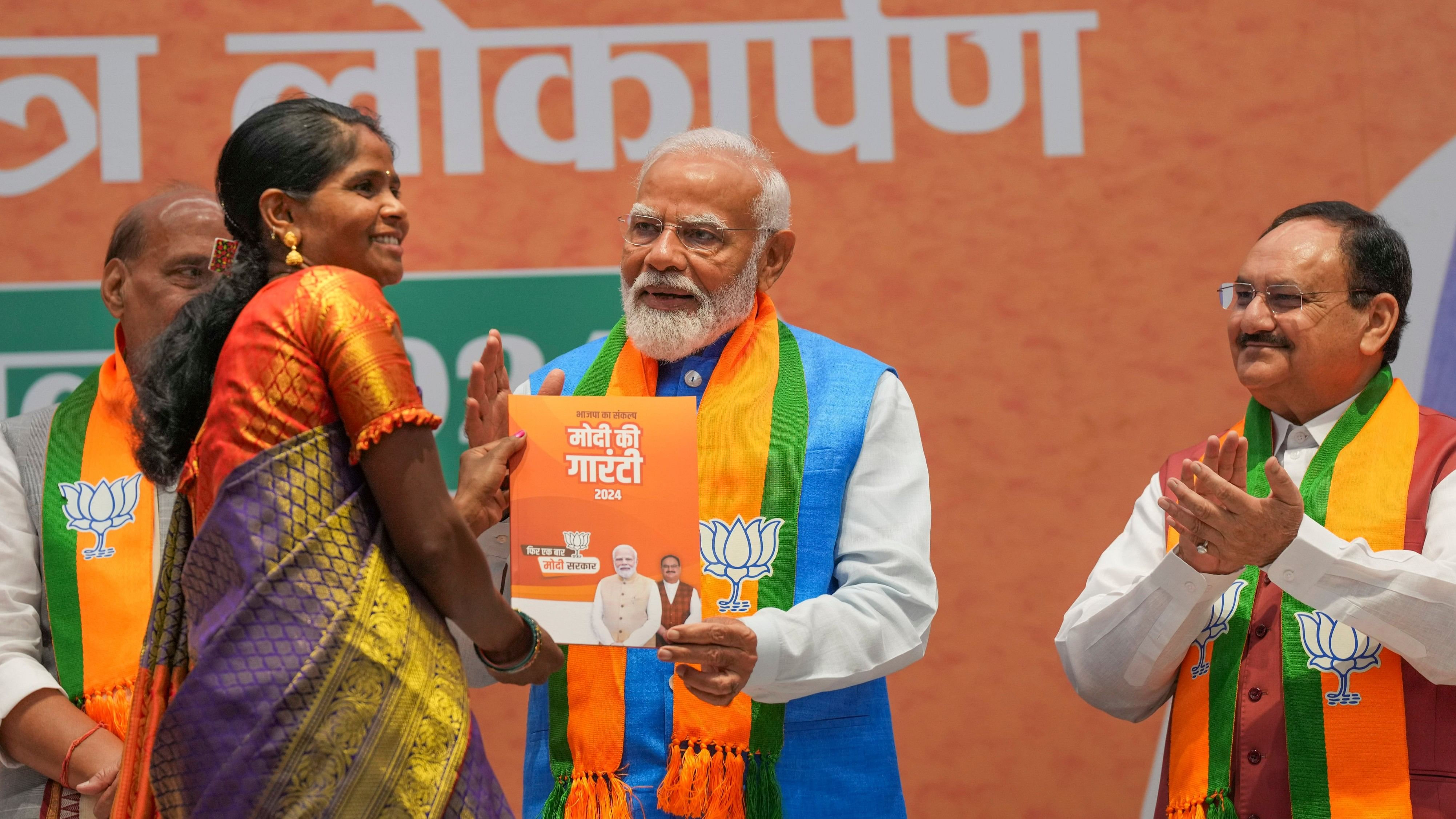 New Delhi: Prime Minister Narendra Modi hands over a copy of BJP's manifesto Modi Ki Guarantee to Lilawati from Bastar who has benefited from the Ujjwala Yojana, at its release for the Lok Sabha polls, at the party headquarters, in New Delhi, Sunday, April 14, 2024. BJP National President JP Nadda is also seen. (PTI Photo/Shahbaz Khan) (PTI04_14_2024_000128B)