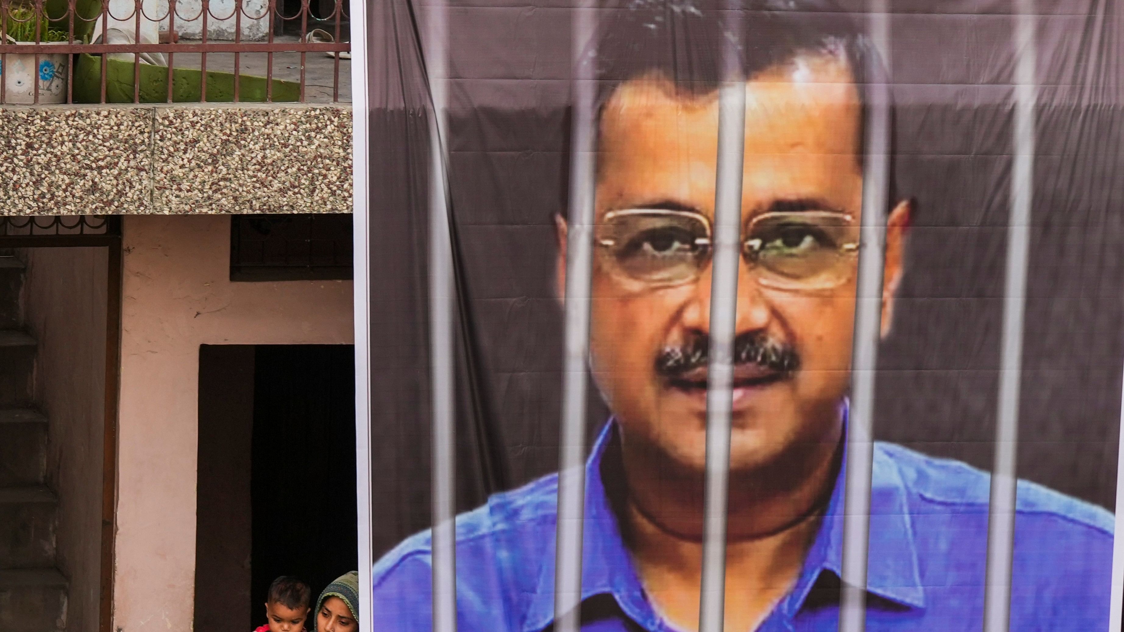 <div class="paragraphs"><p>Locals watch as a large banner of jailed Delhi Chief Minister Arvind Kejriwal hangs from the balcony of a building</p></div>