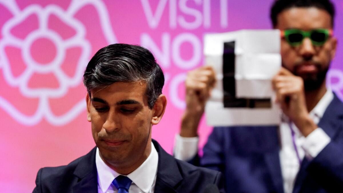 <div class="paragraphs"><p>Independent candidate Niko Omilana holds an "L" sign behind British Prime Minister Rishi Sunak.</p></div>