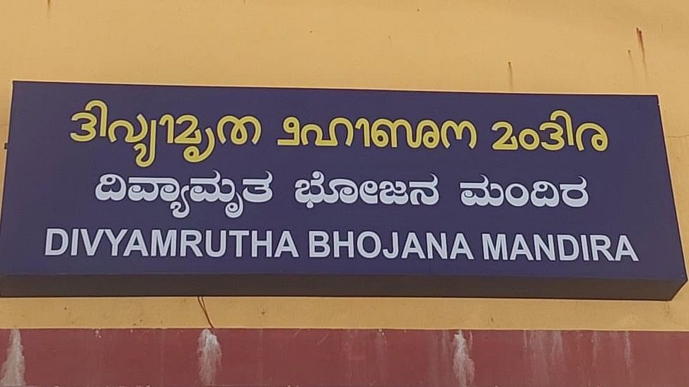 Un letrero de la imagen muestra texto en dos idiomas, con un fondo azul oscuro y letras en amarillo y blanco. En la parte superior, el texto en amarillo está en un idioma dravídico, posiblemente kannada o tulu. Debajo, en letras blancas, aparece el mismo texto en kannada y luego en inglés: "DIVYAMRUTHA BHOJANA MANDIRA". El letrero parece estar ubicado en la pared de un edificio de color beige o amarillo claro.