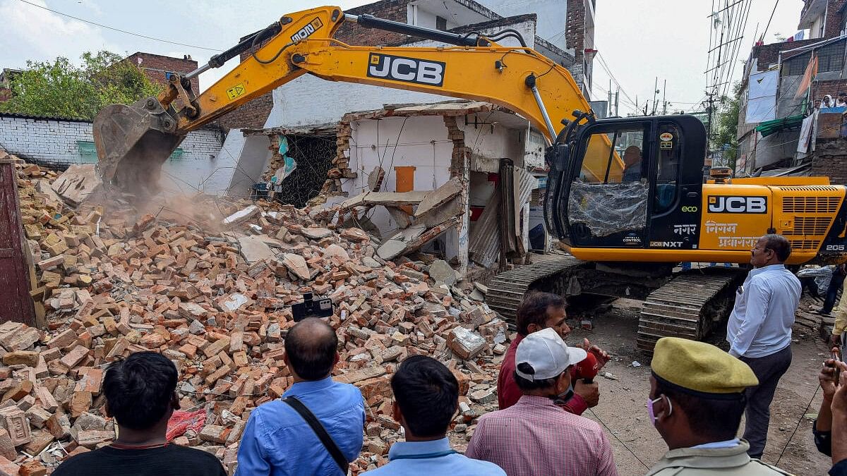 <div class="paragraphs"><p>Police and media watch as a Prayagraj Development Authority bulldozer demolishes properties of Mohammad Gulam, a close aide of Atique Ahmad and accused in Umesh Pal murder case, in Prayagraj.</p></div>