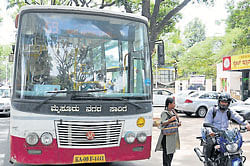 A KSRTC bus stops near a bus shelter on the road in front of TTL College in Saraswatipuram. Cars can be seen parked under the bus shelter. dh photo