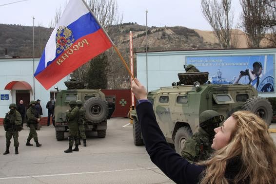A woman waves a Russian flag as armed servicemen wait near Russian military vehicles outside a Ukrainian border guard post in the Crimean town of Balaclava March 1, 2014. Reuters