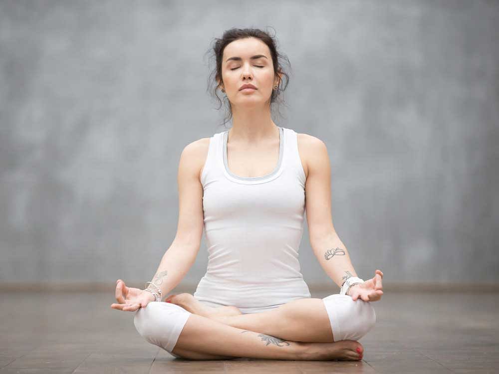 Front view portrait of beautiful young woman with floral tattoos working out against grey wall, resting after doing yoga exercises, sitting in ardha Padmasana, Lotus pose, relaxing. Full length  Breath meditation