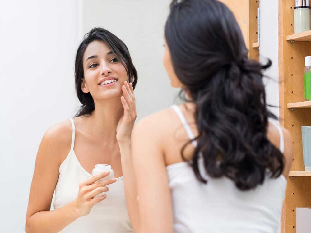 Woman caring of her beautiful skin on the face standing near mirror in the bathroom. Beautiful young woman applying moisturizer on her face. Smiling girl holding little jar of skin cream and applying lotion on face.