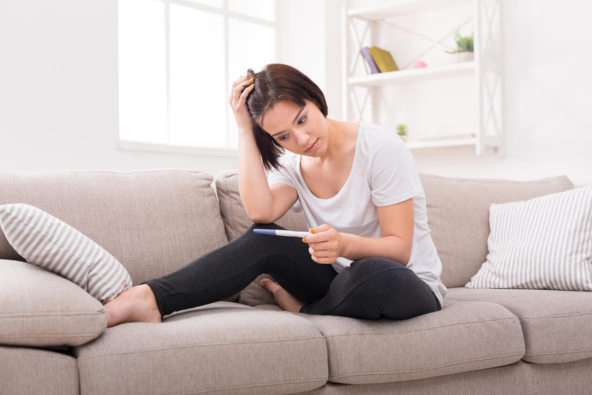 Upset brunette girl checking her recent pregnancy test, sitting on beige couch at home with copy space. Pregnancy, maternity and expectation concept.