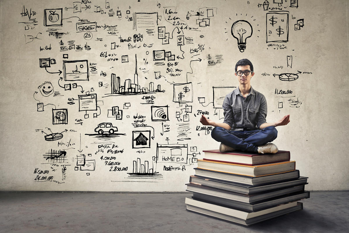 Young Asian guy with nerdy black glasses sitting in yoga position on top of some huge books concentrating in front of a gray wall full of drawings and business concepts.