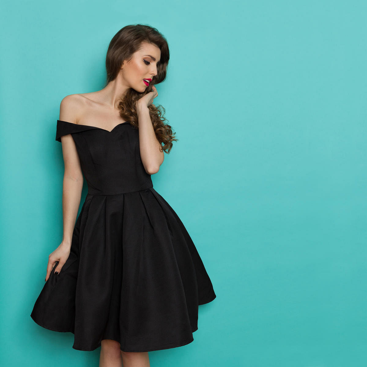 Beautiful young woman in elegant black cocktail dress is looking away. Three quarter length studio shot on turquoise background.