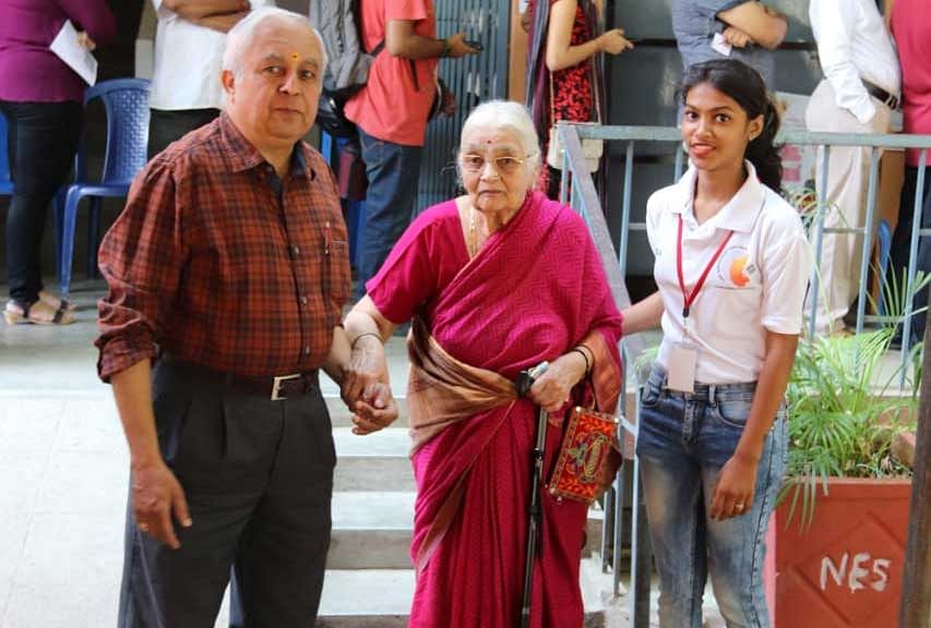 Jayalakshmi, 93, is helped down the steps by her son, Sanjeeva, and NSS volunteer, Prerana Chopra, at the National College, in Basavangudi. (DH Photo)