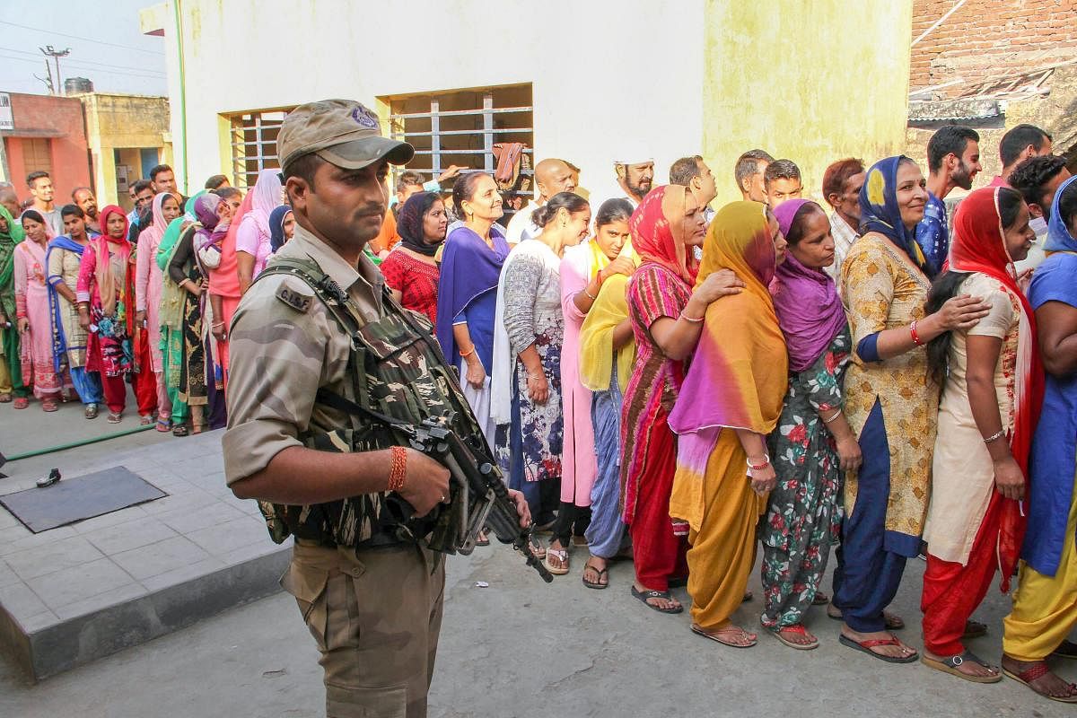 A security person stands guard as people wait in a long queue to cast their votes at a polling station during municipal elections in Bari Brahmana, about 25 kms from Jammu on Saturday. TI