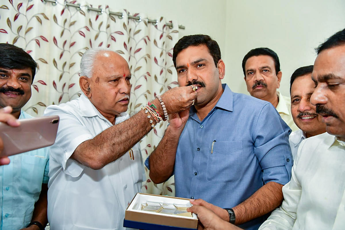 Karnataka Chief Minister BS Yediyurappa offer sweets to BY Vijayendra after BJP won in 12 out of 15 assembly constituencies of Karnataka securing the position of Yediyurappa-led BJP government in the state. (PTI Photo)