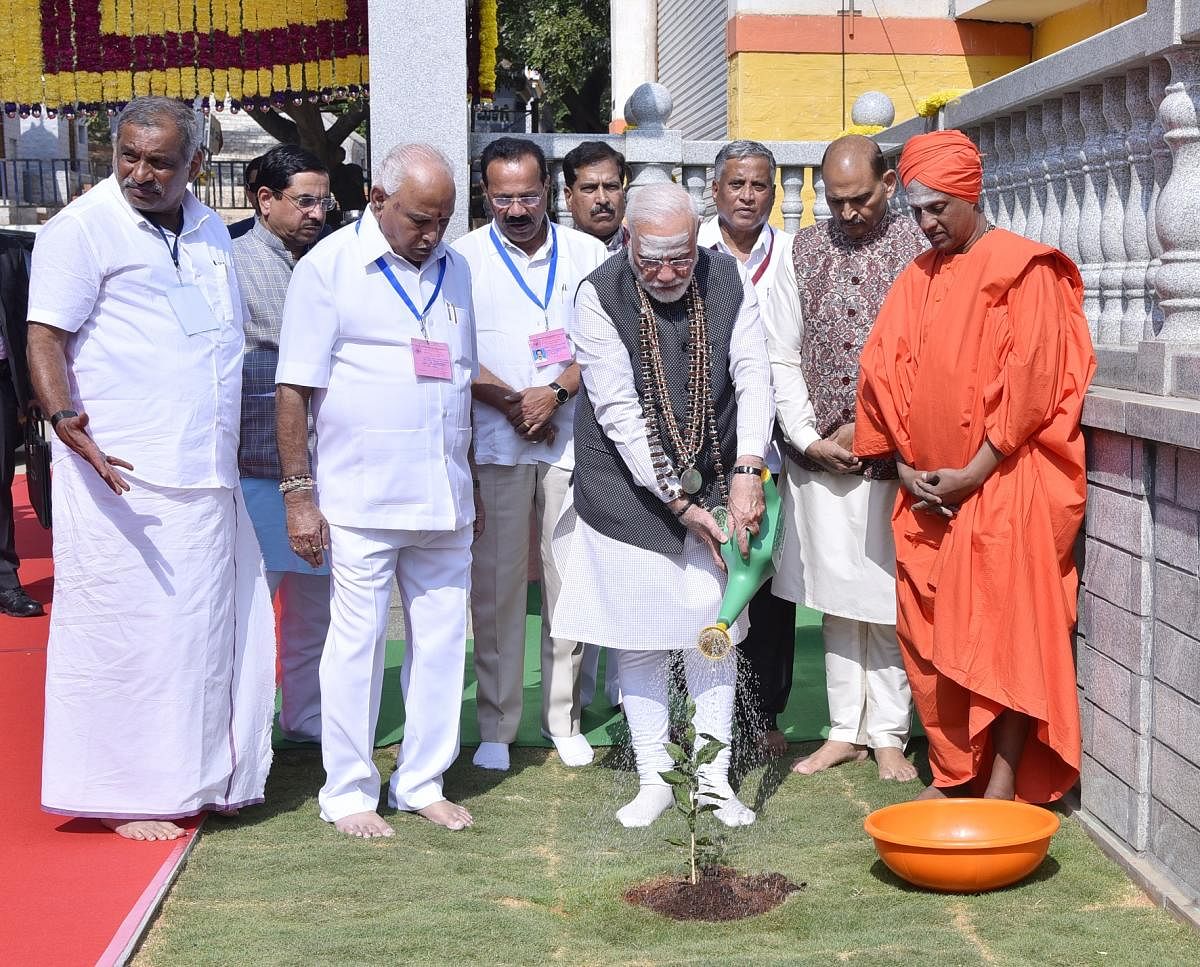 Prime Minister Narendra Modi plants a sapling of Bilva Patre plant on the premises of Shivakumara Swamiji's Gadduge at Siddaganga Mutt in Tumakuru on Thursday. Siddalinga Swamy, Chief Minister B S Yediyurappa, Union ministers Pralhad Joshi, Sadananda Gowd