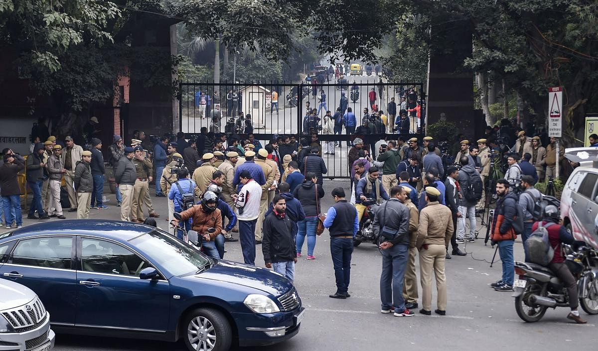 Police personnel guard as students and media are also seen at the main gate of the Jawaharlal Nehru University (JNU), in New Delhi, Monday, Jan. 6, 2020. A group of masked men and women armed with sticks, rods and acid allegedly unleashed violence on the