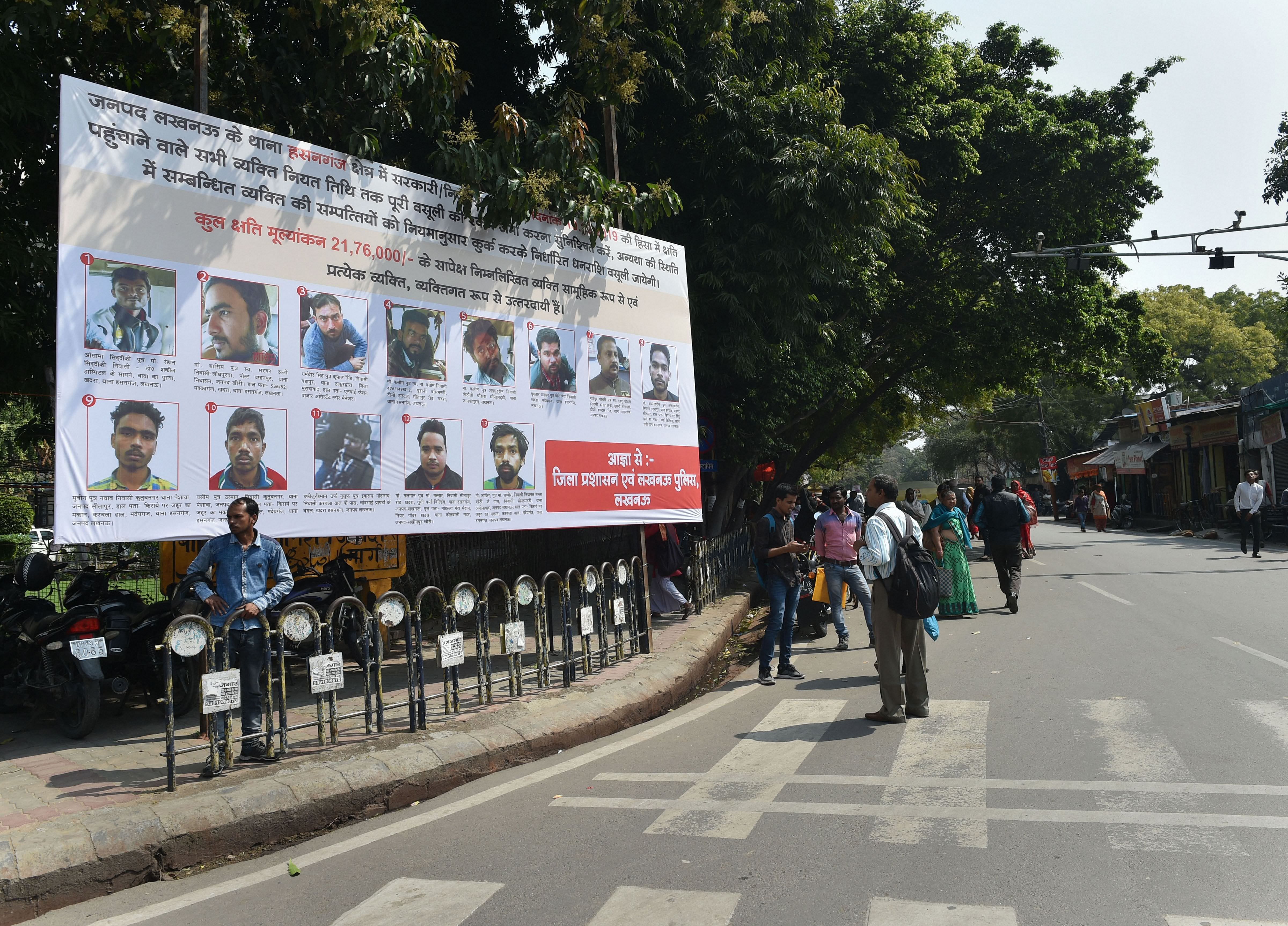 People walk past a poster displaying photographs of those who have been identified to pay the compensation for vandalizing public properties during protests against CAA, in Lucknow. (Credit: PTI)