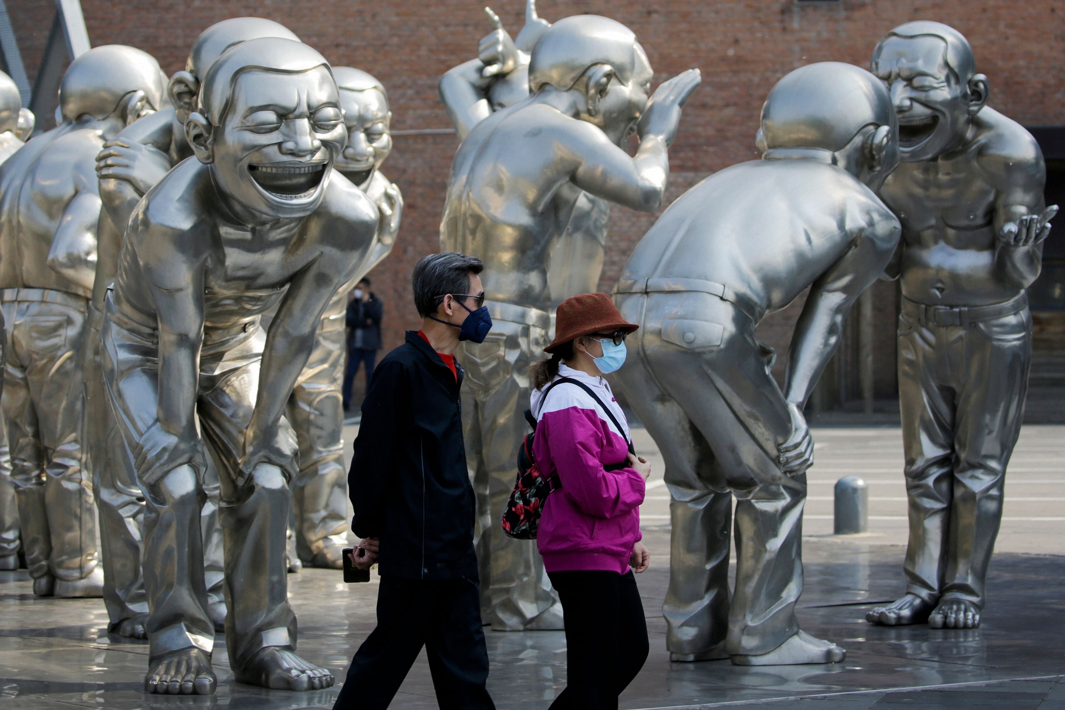 People wearing protective face masks to help curb the spread of the new coronavirus walk by human sculptures on display outside an art gallery in Beijing. (Credit: AP Photo)