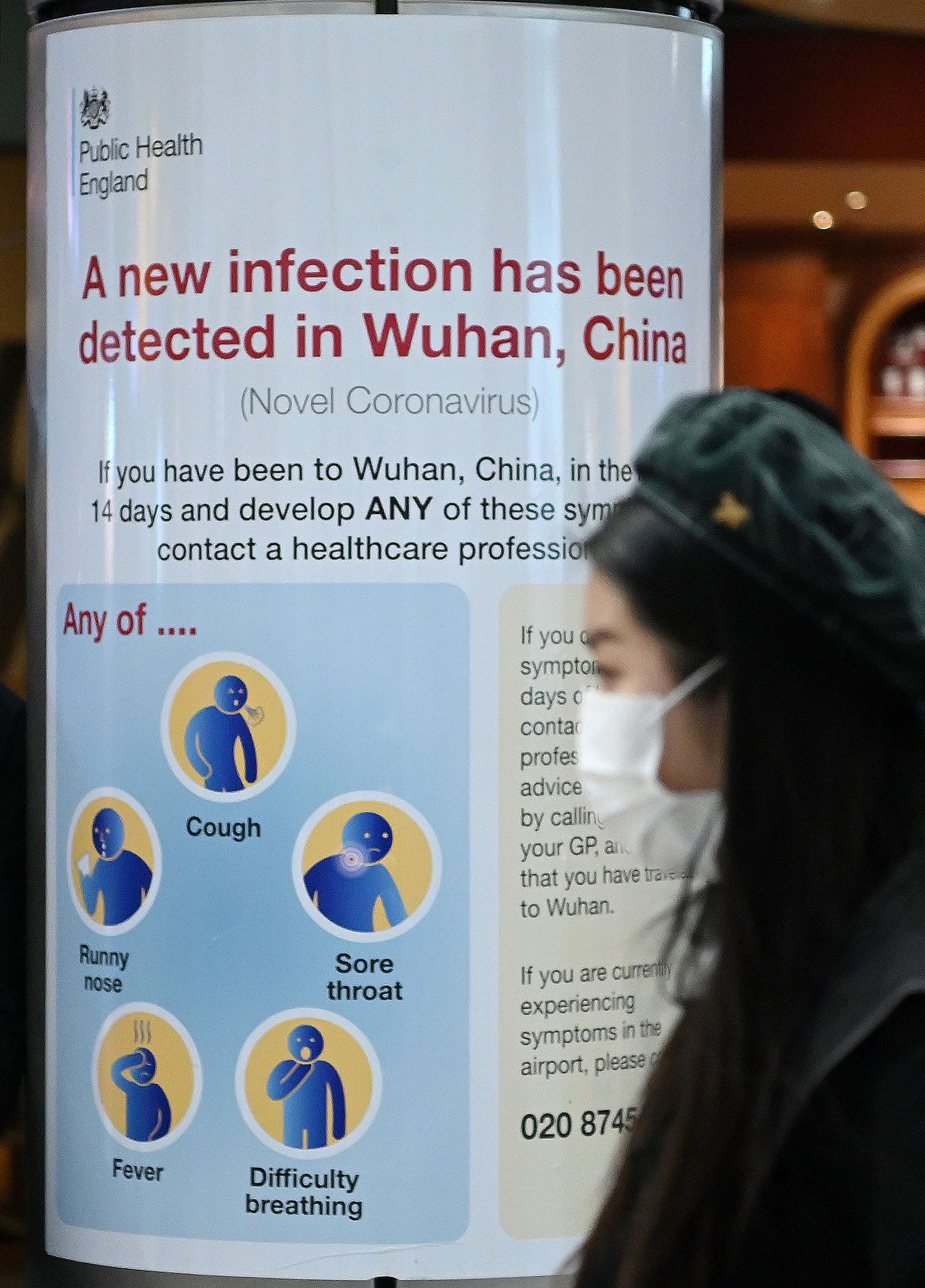 A woman wearing a face mask passes a Public Health England sign, warning passengers arriving on flights into the UK, that a virus, Coronavirus, has been detected in Wuhan in China, at Terminal 4 of London Heathrow Airport in west London on January 28, 2020. (Credit: AFP Photo)