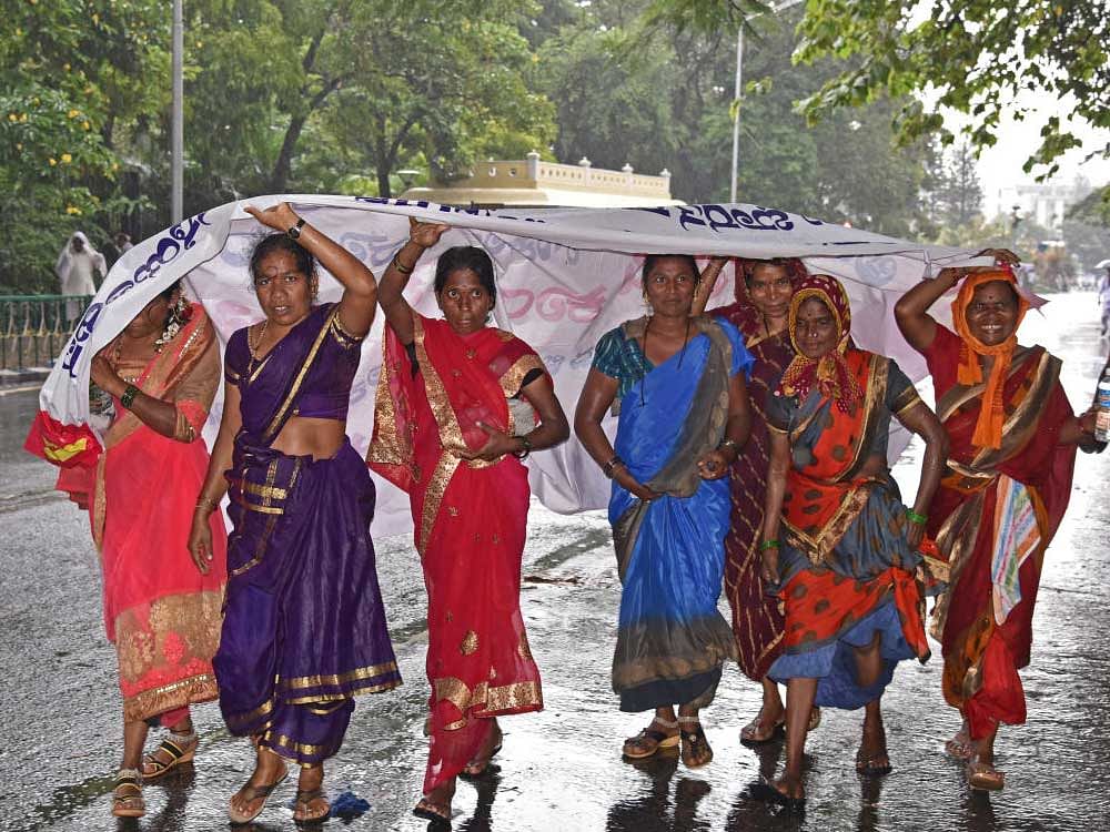 Women who had come for the Valmiki Jayanti celebrations take cover from the rain under a flex banner near Vidhana Soudha on Thursday. DH photo