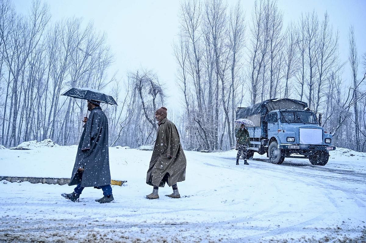 People walk as a security personnel (R) holding an umbrella stands next to a security vehicle near the Dal Lake during a snowfall in Srinagar on January 12, 2020. (AFP Photo)