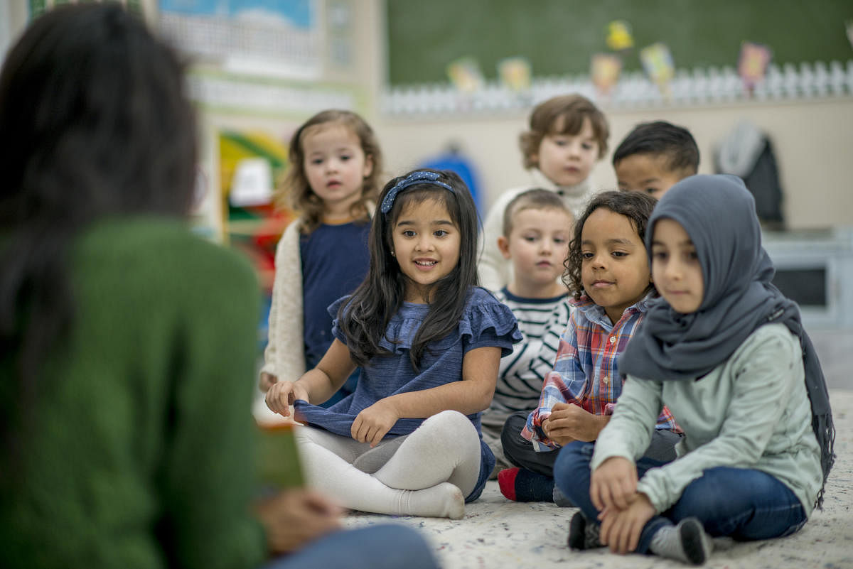 A group of preschool kids are indoors with their teacher. The kids are sitting on the floor and listening to their teacher reading a book.Kids Listening To A Story