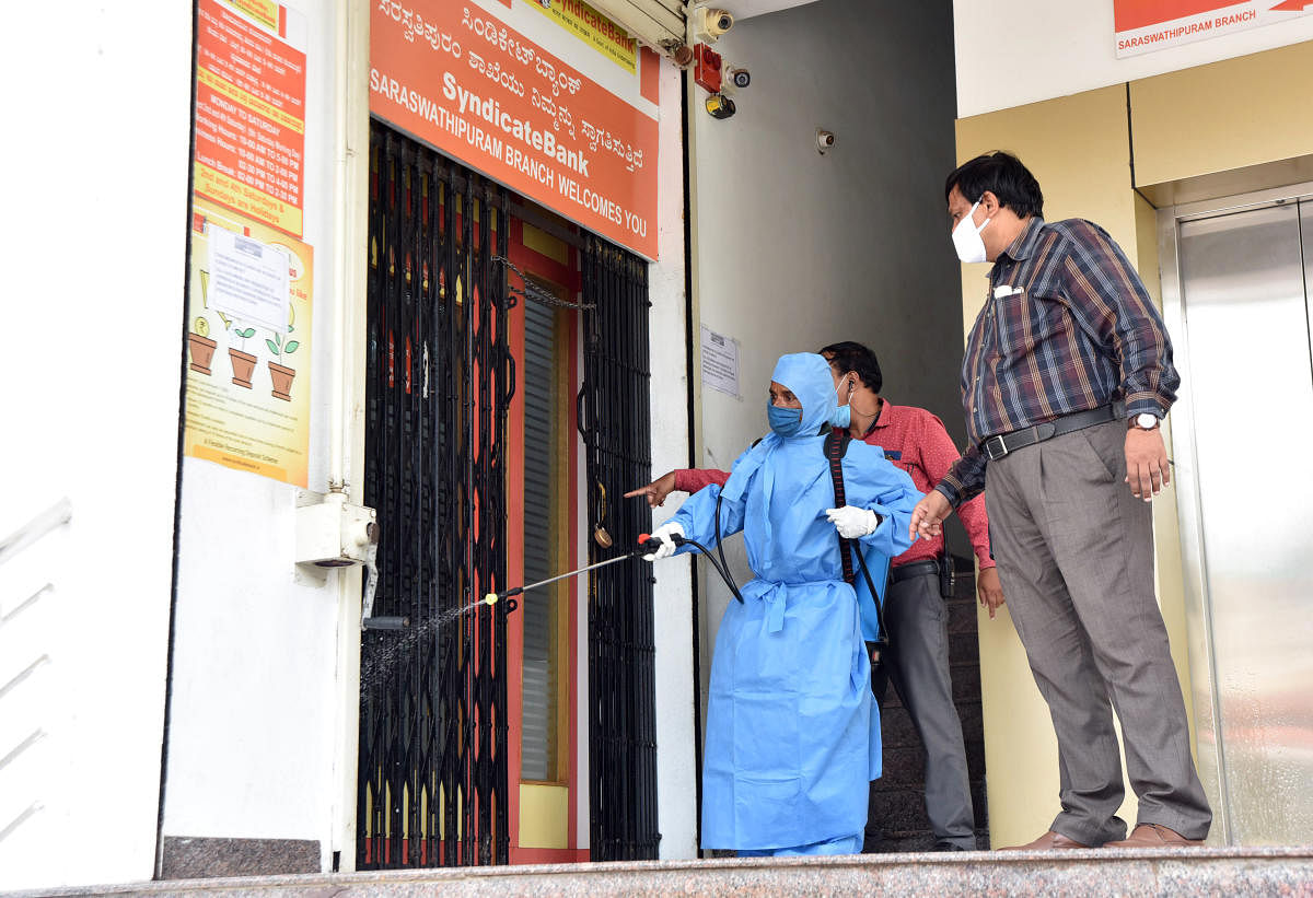 A worker sprays disinfectant at Syndicate Bank branch in Mysuru on Friday. Mysuru City Corporation (MCC) Health Officer Dr Jayanth is seen. DH PHOTO