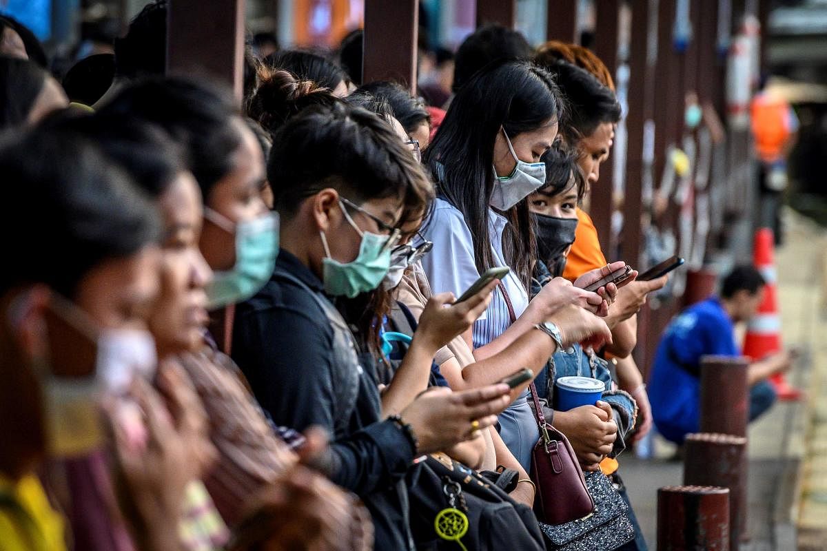 Commuters, wearing facemasks amid fears of the spread of the COVID-19 novel coronavirus, use their mobile phones as they wait for a canal boat in Bangkok on March 2, 2020. Credit/AFP Photo
