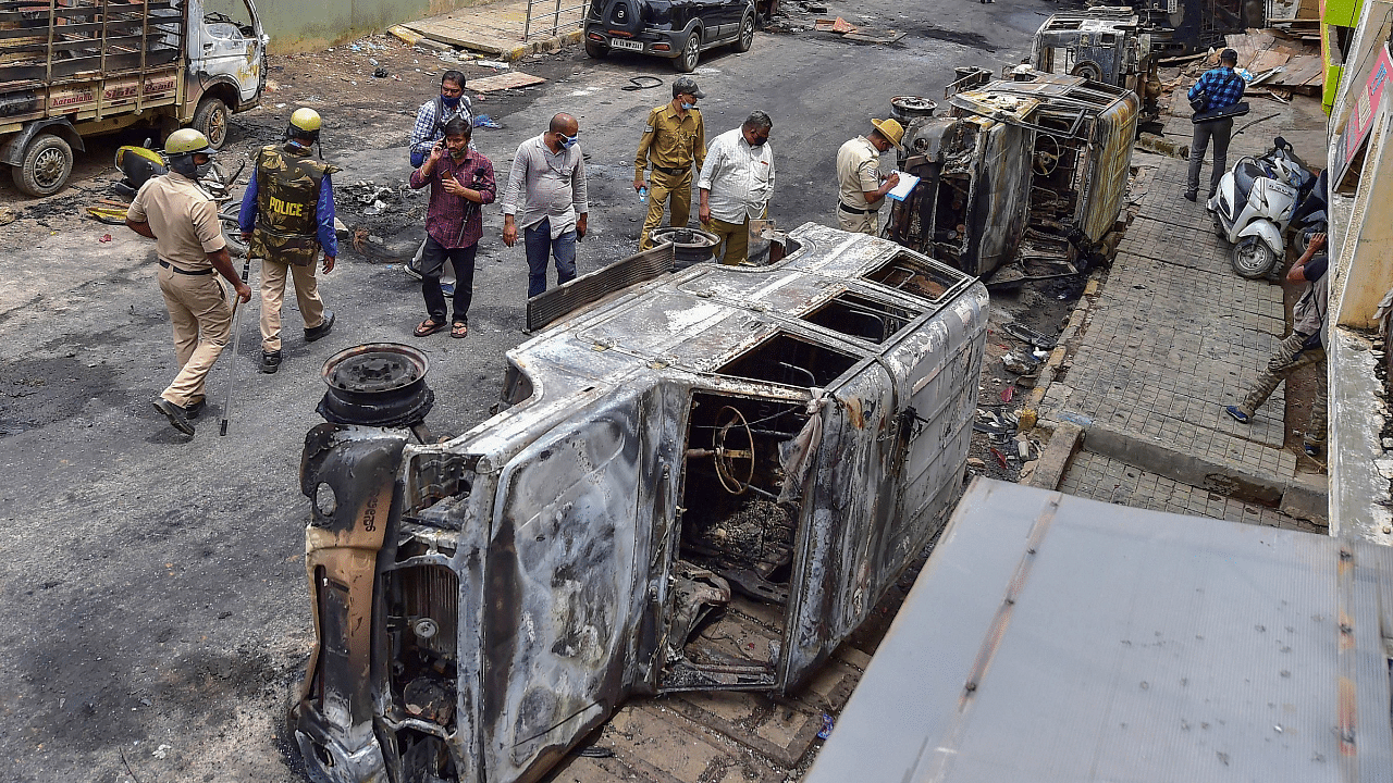 Police and residents walk past charred remains of vehicles vandalised by a mob over a social media post, allegedly by a relative of a Congress MLA, in Bengaluru. Credits: PTI Photo