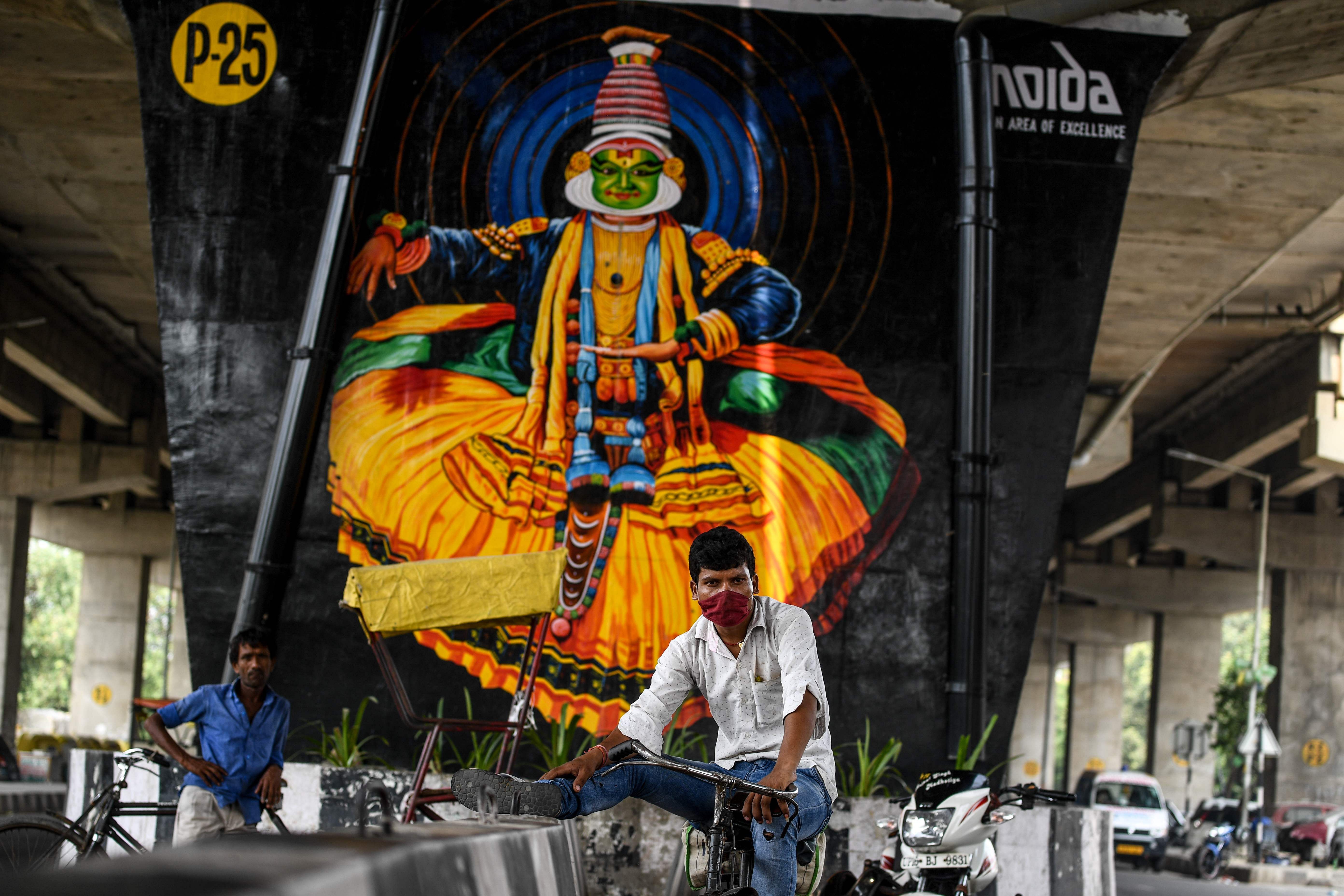 A man sitting on a bicycle takes a break in front of a mural painted on a pillar of a flyover in Noida. Credits: AFP Photo