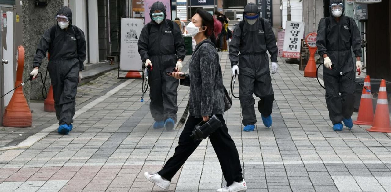 A woman (C) walks past South Korean health officials from Bupyeong-gu Office wearing protective gear spraying disinfectants at a shopping district in Incheon. Credit: AFP