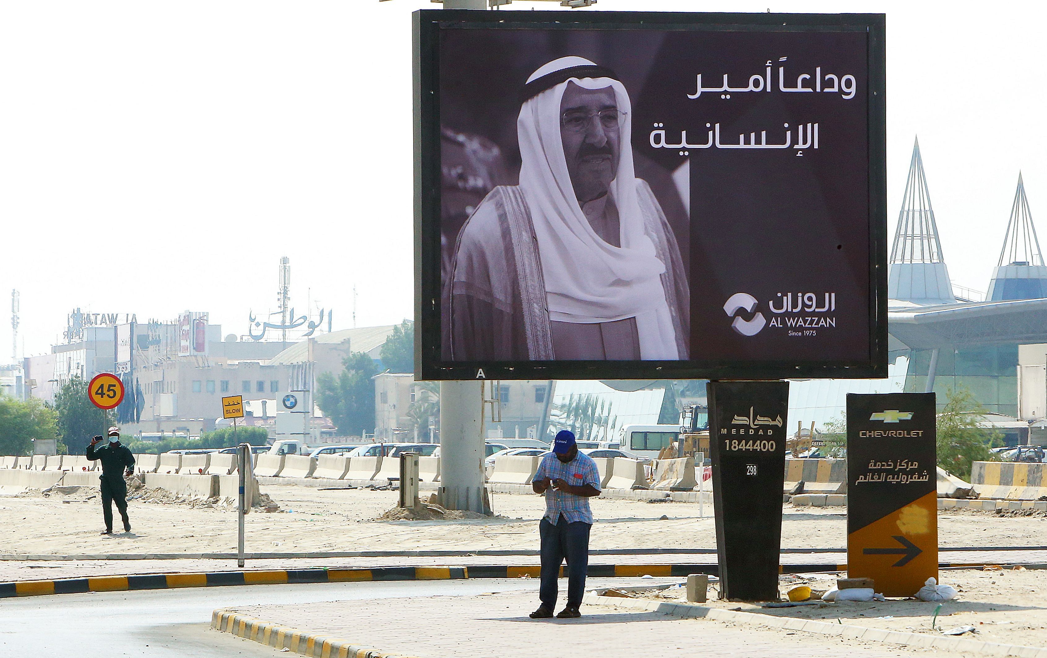 A man stands beneath a billboard displaying the face of Kuwait's late emir Sheikh Sabah al-Ahmad al-Jaber al-Sabah with a message of condolences on his passing in Kuwait City on October 1, 2020. Credit: AFP