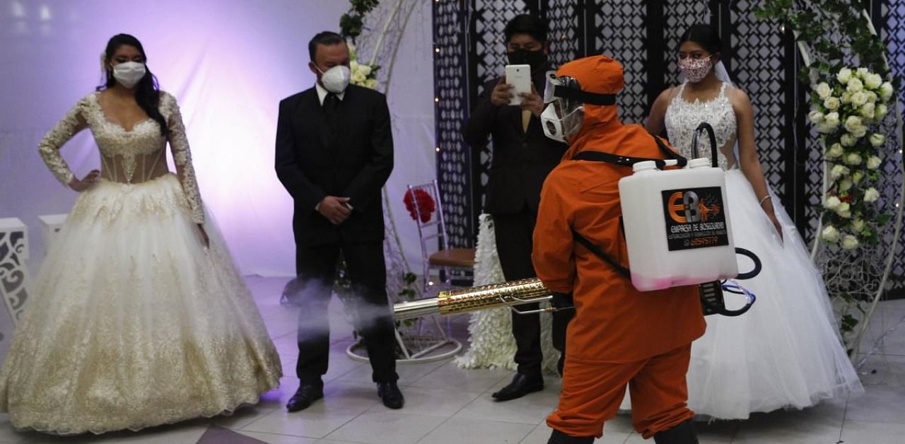 A man wearing a full protection suit sprays disinfectant on two couples in bridal dresses and suits, inside a ballroom after a mock wedding during the partial lifting of restrictions amid the COVID -19 pandemic in La Paz, Bolivia, Wednesday, Oct. 7, 2020. Credit: AP Photo