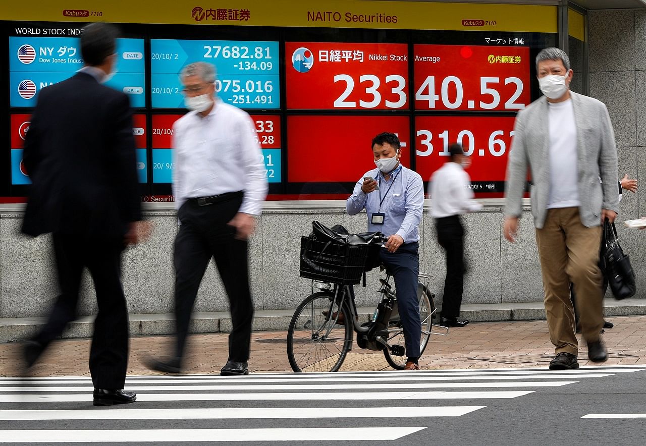 Passersby wearing protective face masks walk past a screen displaying Nikkei share average and world stock indexes. Credits: Reuters Photo