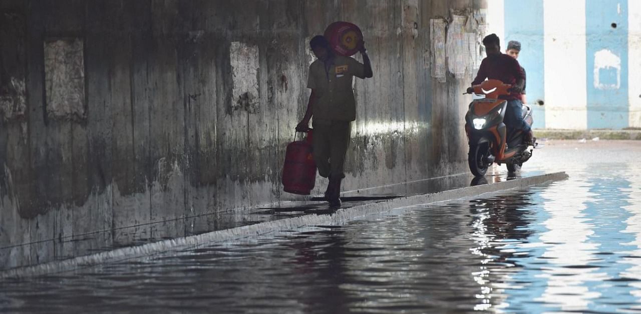 A delivery man carrying gas cylinders walks on a pavement along a waterlogged underpass following heavy rain, in Bengaluru. Credit: PTI